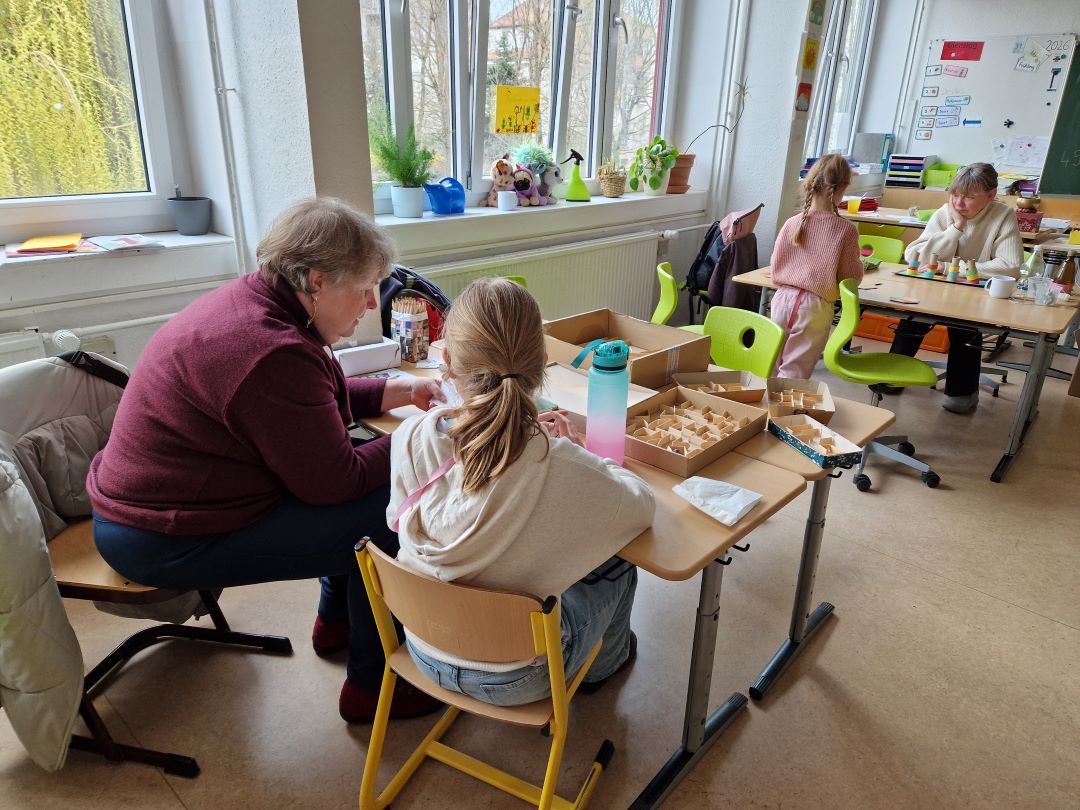 Ein Lehrer sitzt neben einem Schüler an einem Tisch im Klassenzimmer und hilft ihm bei einer Aktivität mit Holzklötzen. Andere Schüler arbeiten an separaten Tischen. Das Klassenzimmer hat große Fenster, Grünpflanzen und Lernposter an den Wänden.