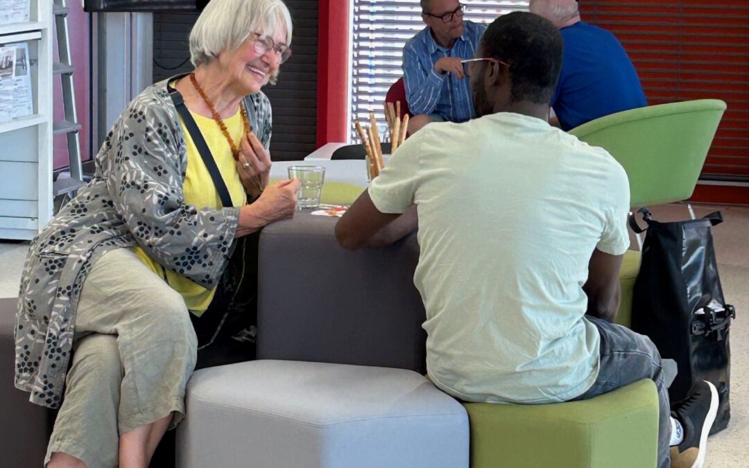 Eine ältere Frau und ein jüngerer Mann sitzen auf bunten Sitzwürfeln, einander zugewandt und unterhalten sich. Im Hintergrund unterhalten sich andere Menschen in einem hell erleuchteten, modernen Innenraum.