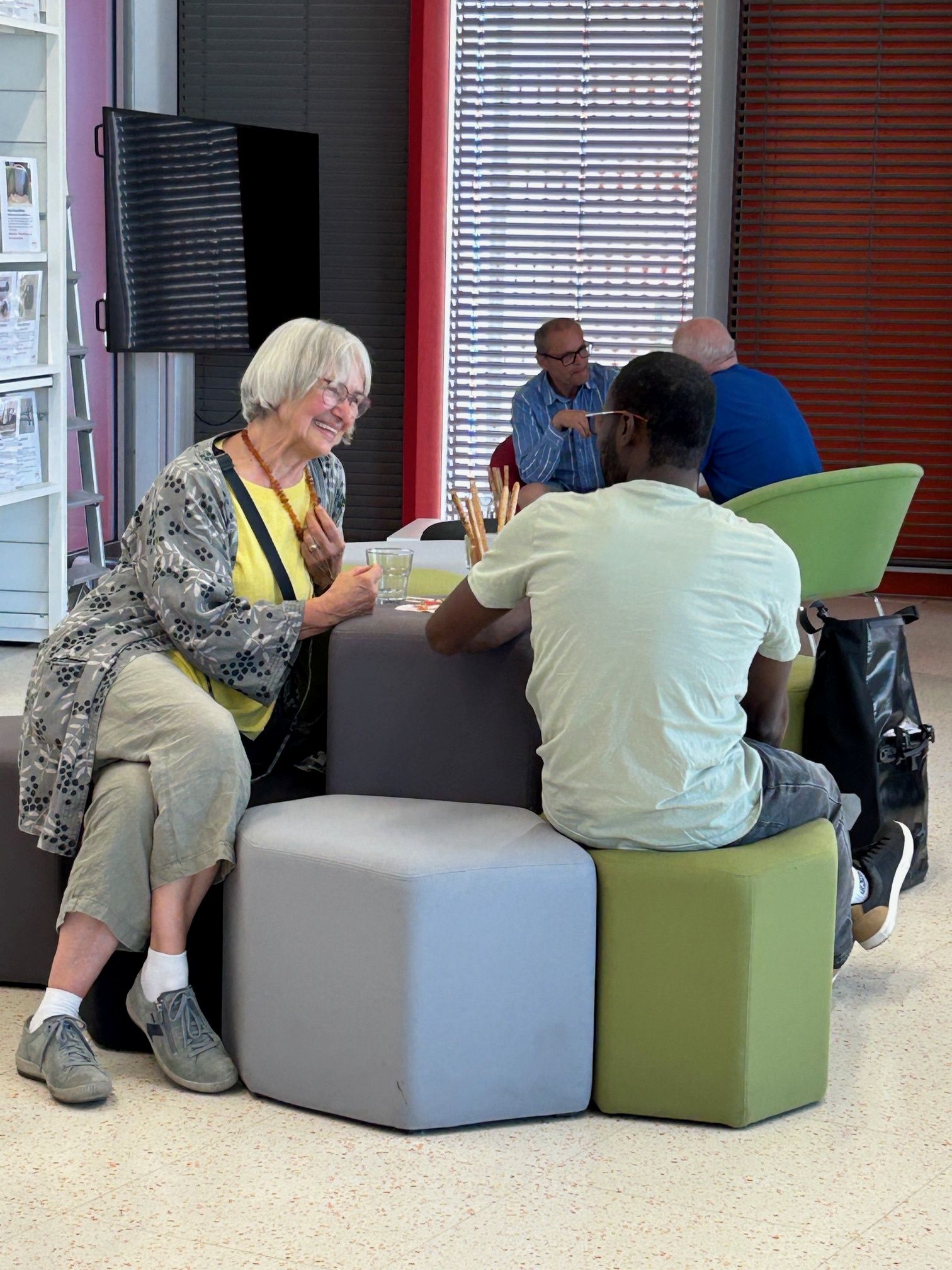 Eine ältere Frau und ein jüngerer Mann sitzen auf bunten Sitzwürfeln, einander zugewandt und unterhalten sich. Im Hintergrund unterhalten sich andere Menschen in einem hell erleuchteten, modernen Innenraum.