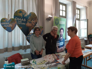 Drei Frauen stehen an einem Informationsstand in einem Innenraum mit Omas for Future-Schildern in Form von Herzen und einem BERR-Banner im Hintergrund. Der Tisch ist mit Broschüren und Flyern bedeckt.