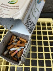 Ein offener Getränkekarton aus Pappe, gefüllt mit gebrauchten Zigarettenstummeln, steht auf einem gelben Metallgitter. Der Karton ist verschmutzt und teilweise beschädigt, mit einem sichtbaren QR-Code und deutschem Text auf der Außenverpackung.