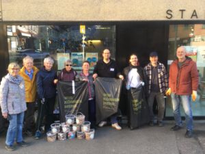 Eine Gruppe von neun Erwachsenen steht vor einem Gebäude und lächelt in die Kamera. Vor ihnen stehen mehrere Eimer mit gesammeltem Müll, und einige Personen halten große schwarze Müllsäcke.
