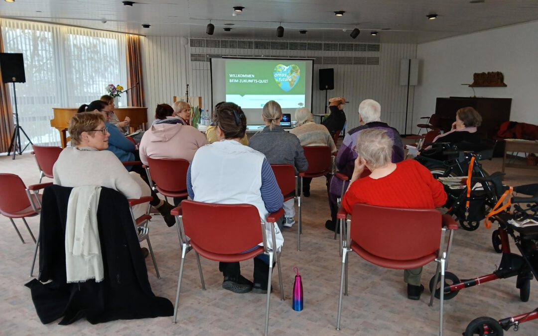 Eine Gruppe von Personen sitzt auf roten Stühlen in einem hellen Raum vor einer Projektionsfläche. Auf der Leinwand wird eine Begrüßungsbotschaft für ein Seniorencafé angezeigt. Einige Teilnehmer benutzen Gehhilfen; Mäntel hängen an den Stühlen in der Nähe.