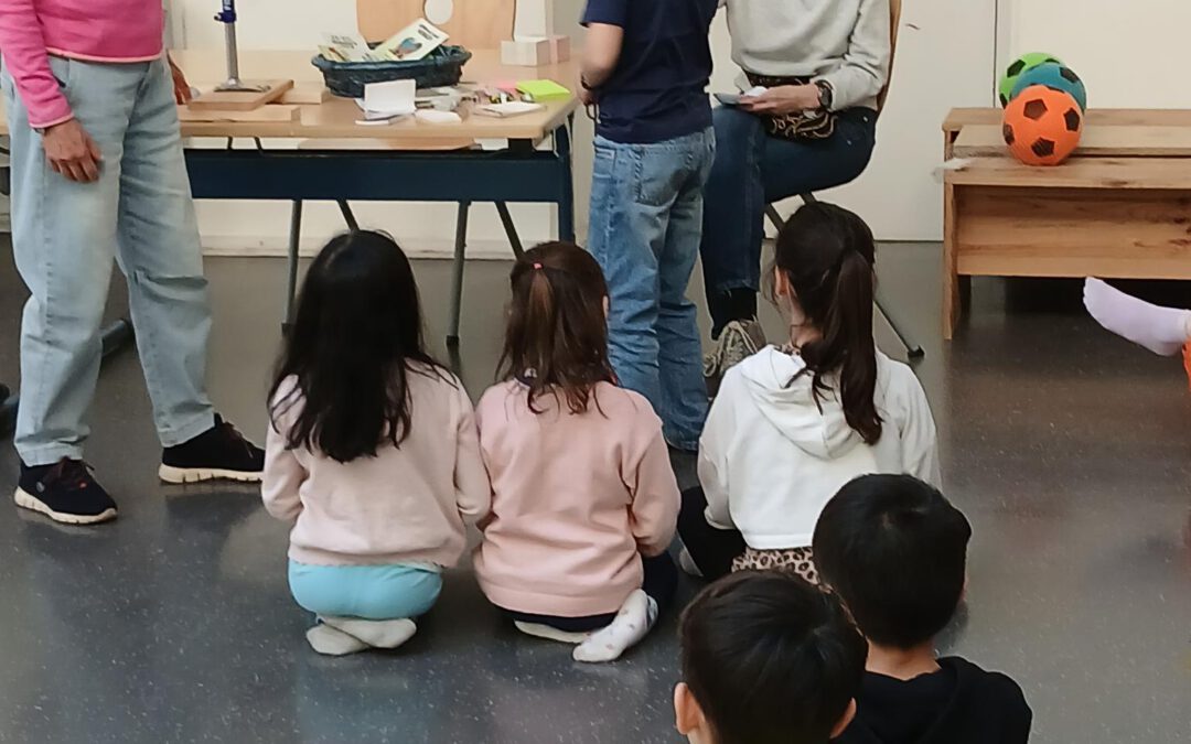Eine Gruppe von Kindern sitzt auf dem Boden vor einem Tisch mit verschiedenen Gegenständen. Zwei Frauen stehen hinter dem Tisch, eine spricht mit einem Jungen, der neben der Gruppe steht. Im Hintergrund sind ein Spinnrad und bunte Bälle zu sehen.