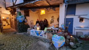 2025 12 Weihnachtsmarkt 1 - Omas for Future Eine Gruppe von Menschen steht hinter einem geschmückten Tisch bei einer Veranstaltung im Freien. Auf Schildern steht "Omas for Future" und auf herzförmigen Postern ist ein Bild der Erde zu sehen. Um den Tisch herum stehen Kerzen, Flugblätter und umweltfreundliche Artikel.