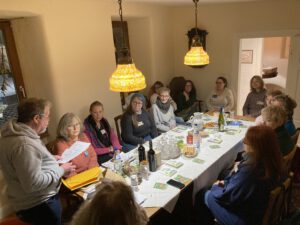 IMG 95481 - Omas for Future Eine Gruppe von Menschen sitzt um einen mit Getränken, Snacks und Papieren gedeckten Esstisch und hört einer Person zu, die steht und spricht. Der Raum ist mit zwei Hängelampen warm beleuchtet.
