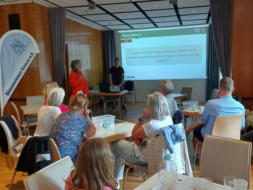 Menschen sitzen an Tischen in einem Konferenzraum und beobachten aufmerksam zwei Moderatoren neben einer Projektionsfläche, die eine Folie mit deutschen Gesundheitstexten zeigt. Auf den Tischen liegen Gläser und Papiere, die auf eine interaktive Quizzen-Sitzung hindeuten.