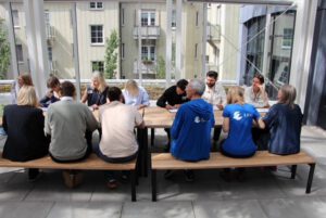 Unterschrift symbolisch© BaumEntscheid Muenchen - Omas for Future Eine Gruppe von Menschen sitzt an einem langen Holztisch im Freien, einige mit dem Gesicht zueinander, andere mit dem Rücken zur Kamera. Zwei Personen tragen blaue Hemden mit weißen Logos. Im Hintergrund sind Gebäude und Grünflächen zu sehen.