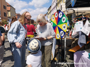 Ehrenamtsfest 2026 2 - Omas for Future Zwei Frauen und ein Kind stehen auf einem Straßenfest im Freien neben einem bunten Spinnrad. Eine Frau spricht mit dem Kind, während sie ein Klemmbrett in der Hand hält. Stände, Menschen und Gebäude säumen die Kopfsteinpflasterstraße.
