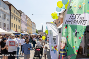 Ein lebhaftes Straßenfest mit farbenfrohen Gebäuden, Ständen und Transparenten mit der Aufschrift „Fest des Ehrenamts“. Unter strahlend blauem Himmel schlendern die Menschen zwischen Ausstellungsständen und Luftballons umher. Logo: Stadt Regensburg.
