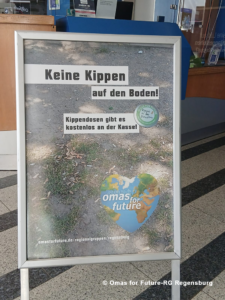 IMG 20260713 WA0000 - Omas for Future Auf einem deutschen Schild steht „Keine Kippen auf den Boden!“, zusammen mit einem Hinweis auf kostenlose Aschenbecher an der Theke und einem „Omas for Future“-Logo in Form eines Herzens mit einer Weltkarte. Das Schild steht auf einem Gehweg im Innenbereich.