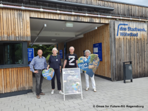 IMG 20260717 WA0001 - Omas for Future Vier ältere Menschen stehen vor dem Stadtwerk Wöhrdbad, halten herzförmige Schilder mit der Aufschrift „Omas for Future“ in den Händen und lächeln. Vor ihnen steht auf einer Staffelei ein Plakat zum Thema Klimaschutz.