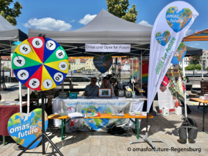 Ein farbenfroher Stand im Freien für „Omas und Opas for Future“ ist mit einem Glücksrad, Bannern und Informationsschildern ausgestattet; an einem sonnigen Tag sitzen zwei Personen hinter dem Tisch.