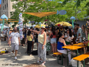 Ein lebhaftes Open-Air-Festival, bei dem sich die Menschen um Tische versammelt haben, sich unterhalten und Getränke genießen. Über ihnen hängen bunte Fischdekorationen. Eine Frau mit Sonnenbrille und Strohhut posiert und zeigt auf etwas, während sich andere in der sonnigen Kulisse unter die Gäste mischen.