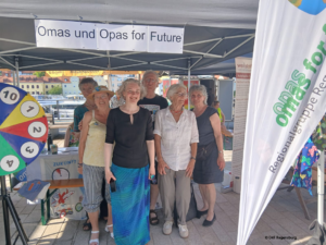 Nachhaltigkeitsfest 2026 3 - Omas for Future Eine Gruppe älterer Menschen steht lächelnd unter einem Zelt, an dem ein Banner mit der Aufschrift „Omas und Opas für die Zukunft“ hängt. Auf den Tischen um sie herum liegen bei einer Veranstaltung im Freien ein buntes Glücksrad und Informationsmaterialien.
