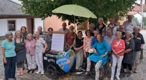 2026 08 Gruppentreffen Disibodenberg2 scaled e1788177639264 - Omas for Future Eine Gruppe älterer Menschen posiert im Freien neben einem Fahrrad mit einem grünen Regenschirm und einem Schild mit der Aufschrift „Omas for Future“. Die Gruppe lächelt vor einem großen Baum und Gebäuden im Hintergrund.