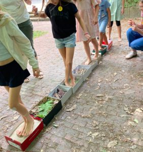 2026 Projekttage 1 Biberach O4F - Omas for Future Im Rahmen einer sensorischen Aktivität im Freien laufen Kinder barfuß durch Kisten, die mit verschiedenen Naturmaterialien wie Blättern, Zweigen und Steinen gefüllt sind. Ein Erwachsener beobachtet sie aus der Nähe von einem gepflasterten Weg aus, der von Blättern umgeben ist.