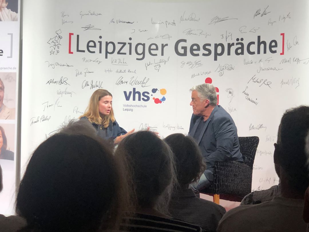Auf einer Bühne im Rahmen der Veranstaltung „Leipziger Gespräche“ sitzen zwei Personen und unterhalten sich. Im Hintergrund sind zahlreiche Unterschriften und das Logo der vhs Leipzig zu sehen. Im Vordergrund sind Zuschauer zu sehen.