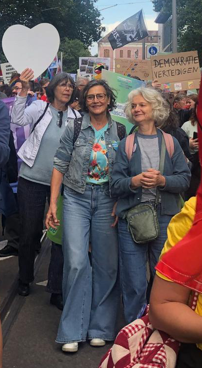 Eine Gruppe von Menschen nimmt an einer Straßenkundgebung teil. Im Vordergrund lächeln drei Frauen; eine von ihnen hält ein herzförmiges Schild in der Hand. Im Hintergrund sind verschiedene Transparente und Schilder zu sehen.