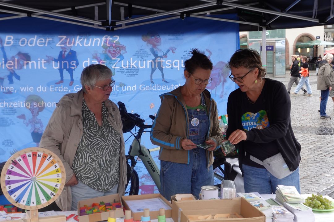 Drei Frauen stehen an einem Stand im Freien, auf dem ein buntes Spinnrad, Broschüren und ein blaues Transparent zum Thema Zukunft zu sehen sind. Zwei Frauen schauen sich Unterlagen an, während die dritte gestikuliert. Im Hintergrund sind Menschen zu sehen, die vorbeigehen.
