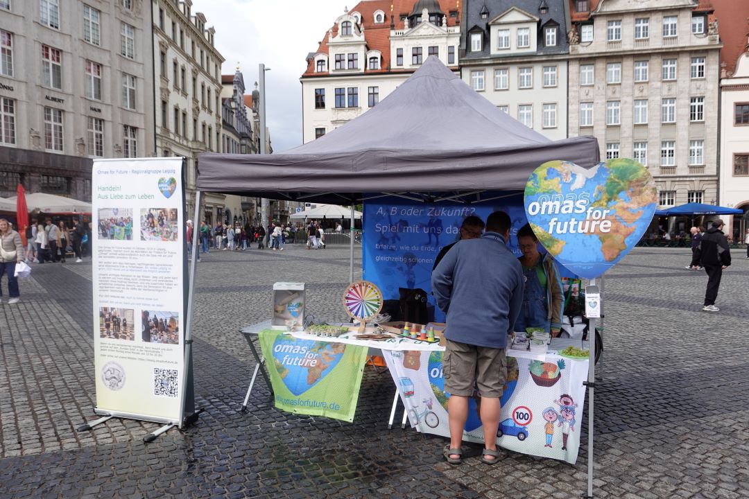 Auf einem Stadtplatz wirbt ein Stand mit einem grauen Vordach für „Omas for Future“. Zwei Personen stehen am Stand, auf dem Broschüren, ein Spinnrad und Plakate ausliegen. Im Hintergrund sind historische Gebäude zu sehen.