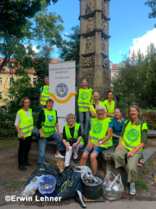 Eine Gruppe von Menschen in gelben Westen posiert vor einem Denkmal mit Säcken voller eingesammelter Abfälle. Hinter ihnen ist ein Banner von Soroptimist International Deutschland zu sehen. Einige sitzen, andere stehen, und das Ganze spielt sich an einem sonnigen Tag im Freien ab.