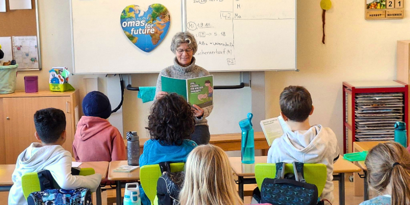 Eine ältere Frau liest einer Gruppe kleiner Kinder, die mit gekreuzten Beinen auf einer blauen Matte sitzen und ihr in einem farbenfrohen Klassenzimmer gegenüberstehen, aus einem Bilderbuch vor.