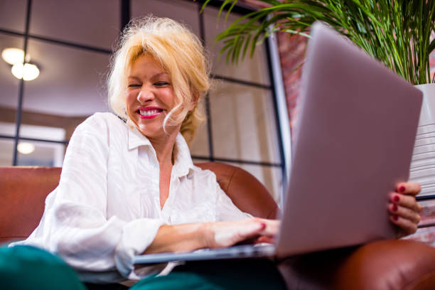 Eine lächelnde Frau mit blonden Haaren sitzt auf einem braunen Sofa und arbeitet an einem Laptop in einem modernen, hell beleuchteten Raum mit großen Fenstern und einer Topfpflanze im Hintergrund.