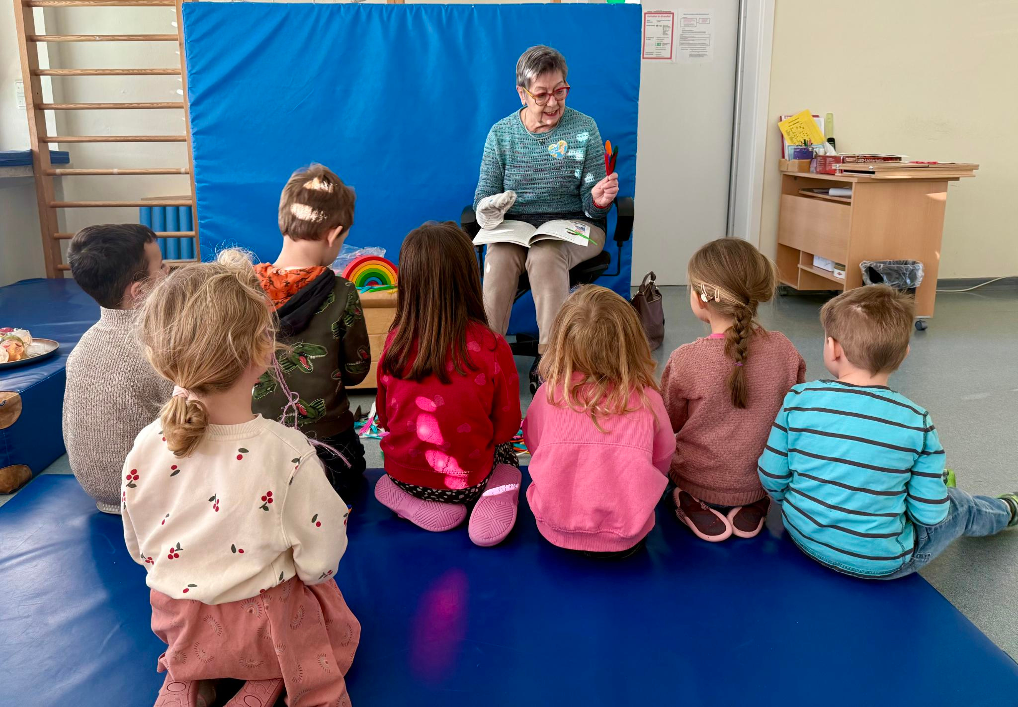 Eine ältere Frau liest einer Gruppe kleiner Kinder, die mit gekreuzten Beinen auf einer blauen Matte sitzen und ihr in einem farbenfrohen Klassenzimmer gegenüberstehen, aus einem Bilderbuch vor.