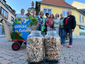 2026 09 Kippensammeln 2 - Omas for Future Eine Gruppe älterer Menschen steht auf einem Stadtplatz hinter einem Wagen mit einem Schild Omas for future heart, im Vordergrund stehen zwei Gläser voller Zigarettenkippen.