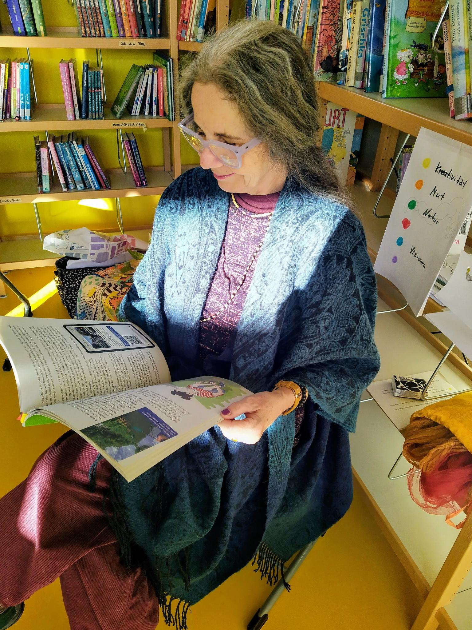 Eine Frau mit langen grauen Haaren und einer Brille sitzt in einer Bibliothek und liest in einem aufgeschlagenen Buch.