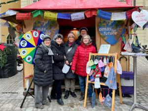 O4F Lucrezia Markt 2025 - Omas for Future Fünf ältere Erwachsene in Winterkleidung stehen lächelnd vor einem hölzernen Marktstand in Regensburg, der mit "Omas for Future"-Schildern, einem bunten Glücksrad und handgefertigten Stoffen geschmückt ist, die zum Verkauf angeboten werden.