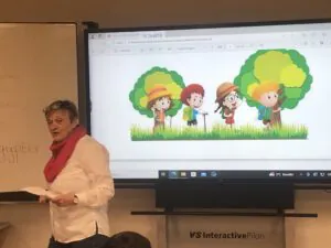Eine Frau steht vor einem Klassenzimmer neben einem Bildschirm, auf dem ein Zeichentrickbild von vier Kindern mit Hüten zwischen Bäumen zu sehen ist. Die Frau hält Papiere in der Hand und scheint vor der Klasse zu sprechen.