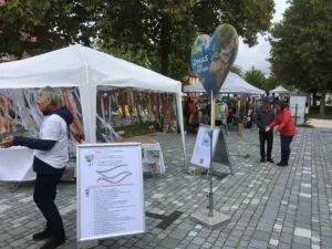 Veranstaltung im Freien mit weißen Zelten, Informationspostern und bunten Bändern. Die Menschen laufen, reden und tauschen sich aus. Ein großes herzförmiges Schild mit einer Weltkugel und dem Text "Omas for Future" steht an prominenter Stelle.
