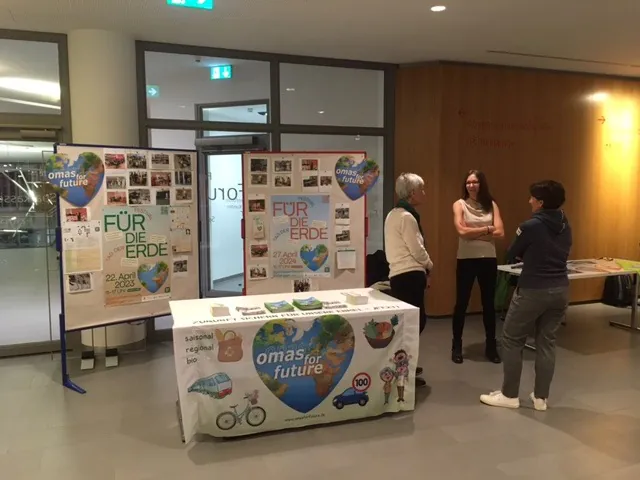 Ein Informationsstand von Omas for Future zeigt Plakate und Flugblätter zum Thema Klimaschutz. Drei Personen unterhalten sich neben dem Tisch, der mit umweltfreundlichen Bildern und Slogans dekoriert ist. Der Stand befindet sich in einem Innenraum mit heller Beleuchtung.