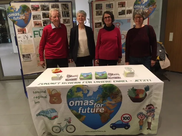 Vier ältere Erwachsene stehen hinter einem Tisch mit einem "Omas for Future"-Transparent und legen Broschüren und Obst aus. Hinter ihnen sind Plakate über Nachhaltigkeit und die Organisation zu sehen.