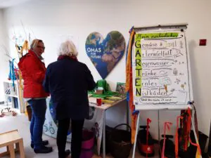 signal 2024 04 20 13 37 49 499 2 - Omas for Future Zwei Frauen stehen in einem Raum neben einem großen Flipchart mit bunten Zeichnungen und deutschem Text über Gartenarbeit. Ein herzförmiges "Omas for Future"-Schild hängt an der Wand. Verschiedene Unterrichtsmaterialien und Zubehör sind zu sehen.