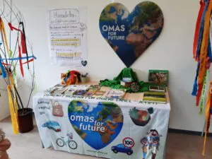 Ein Schautisch mit dem Logo von Omas for Future, bedeckt mit Büchern, Broschüren und umweltfreundlichen Artikeln. Oben an der Wand hängt ein herzförmiges Poster der Erde, links ein Schild mit Informationen zum Klimaschutz.