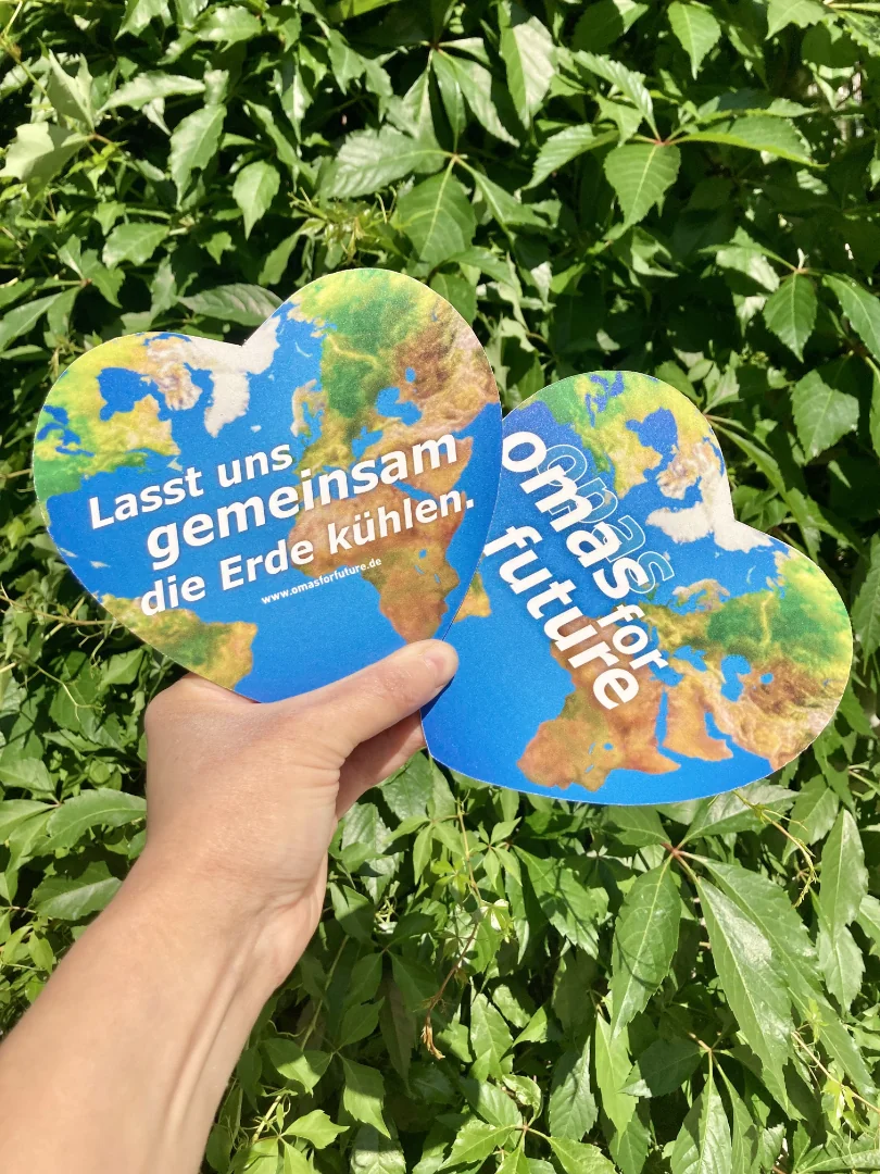 Eine Hand hält zwei herzförmige Ausschnitte mit Bildern der Erde. Auf einem steht Lasst uns gemeinsam die Erde kühlen, auf dem anderen omas für die Zukunft. Grüne Blätter füllen den Hintergrund.
