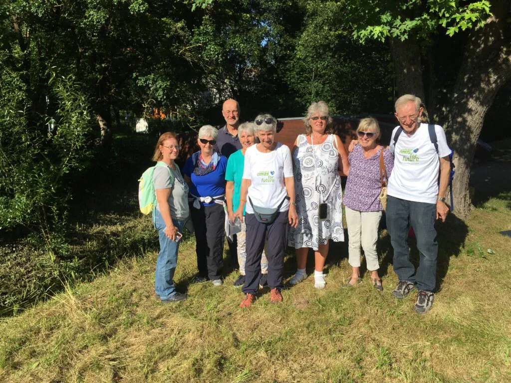 Eine Gruppe von acht älteren Erwachsenen, von denen einige eine Sonnenbrille tragen, posieren an einem sonnigen Tag im Freien zusammen. Sie stehen im Gras in der Nähe von Bäumen und lächeln in die Kamera. Zwei Personen tragen weiße T-Shirts mit grüner Aufschrift.