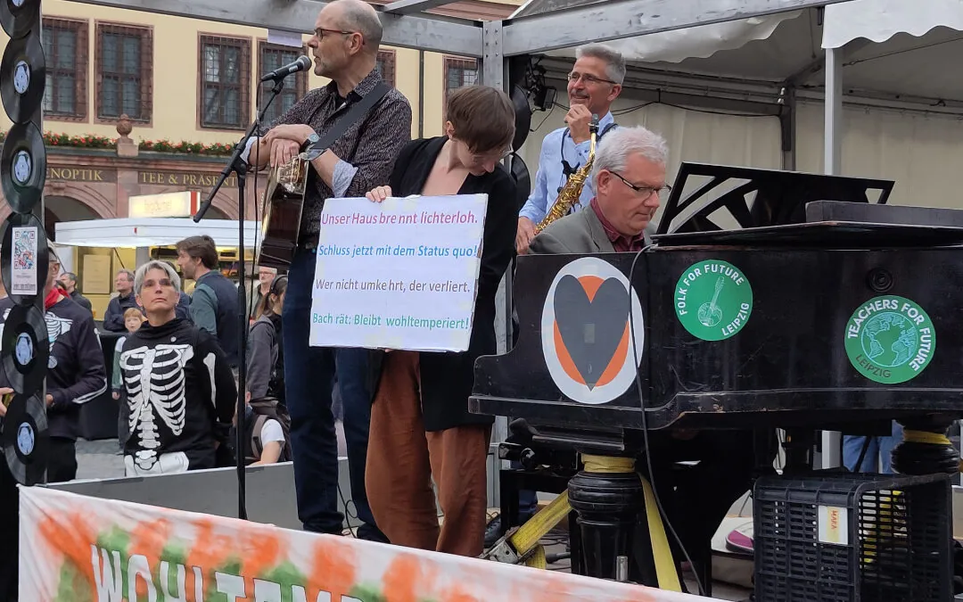 Eine Band tritt auf einer Freilichtbühne auf. Eine Person hält ein Schild in deutscher Sprache, während andere Instrumente spielen, darunter ein Klavier und ein Saxophon. Auf dem Klavier sind Fridays for Future-Aufkleber zu sehen. Im Hintergrund sieht ein Publikum zu.