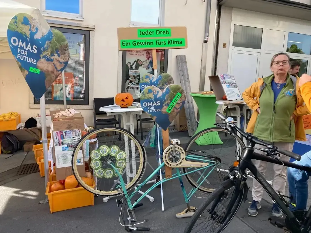 Ein Klima-Aktionsstand mit Omas for Future-Schildern, Kürbissen, einem geschmückten Fahrrad, Displays mit Klimathemen und einer Frau in einer gelben Jacke, die neben dem Stand steht. Auf einem Schild steht "Jeder Dreh, ein Gewinn fürs Klima".