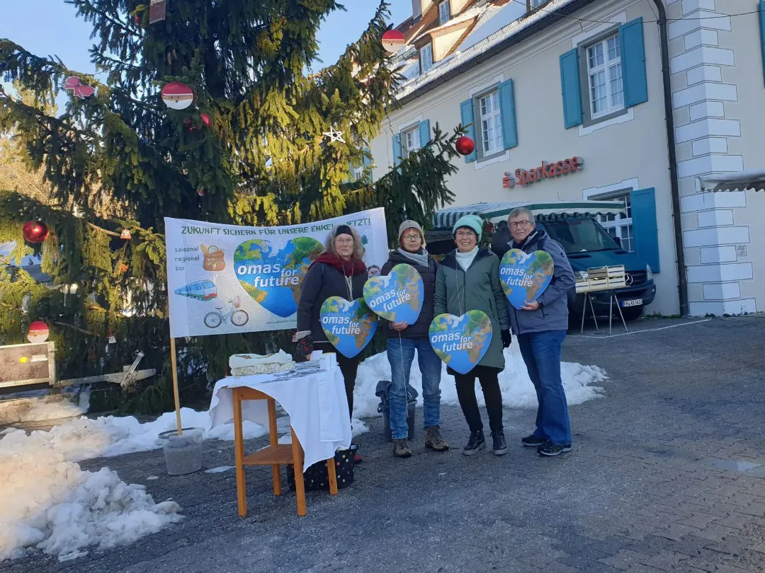 Vier Personen stehen draußen an einem großen Weihnachtsbaum und einem Tisch und halten bunte "omas for future"-Schilder in die Höhe. Hinter ihnen hängt ein Transparent vor einem weißen Gebäude mit blauen Fensterläden. Auf dem Boden liegt Schnee.