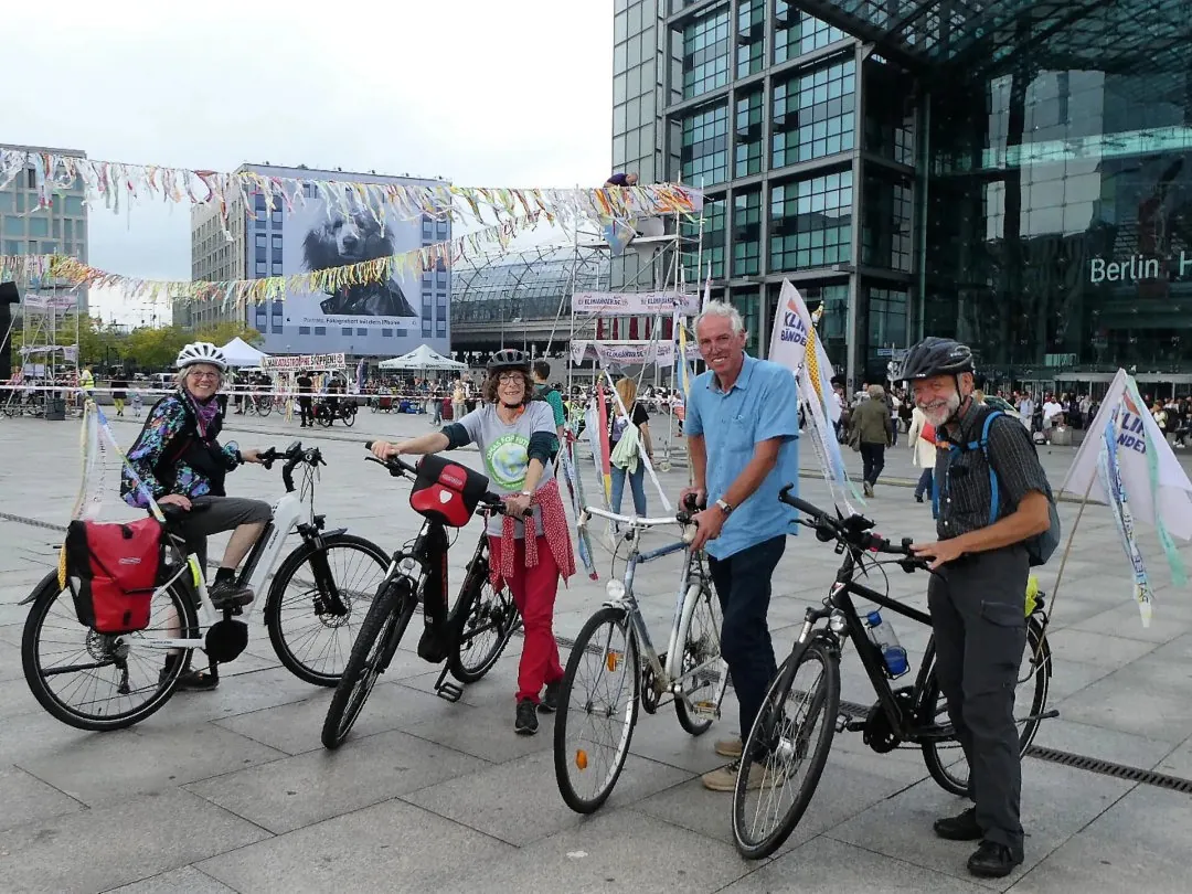 Vier lächelnde Erwachsene mit Fahrrädern posieren auf einem mit bunten Luftschlangen geschmückten städtischen Platz. Im Hintergrund sind Gebäude und Menschen zu sehen, mit dem Schild "Berlin Hbf" an einem Glasgebäude hinter ihnen.
