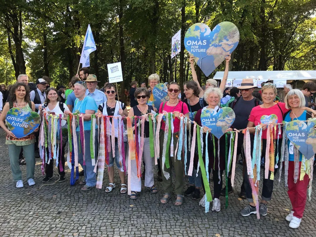 Eine Gruppe älterer Menschen steht im Freien und hält bunte Stoffbänder und herzförmige Schilder mit der Aufschrift OMAS FÜR DIE ZUKUNFT in die Höhe.