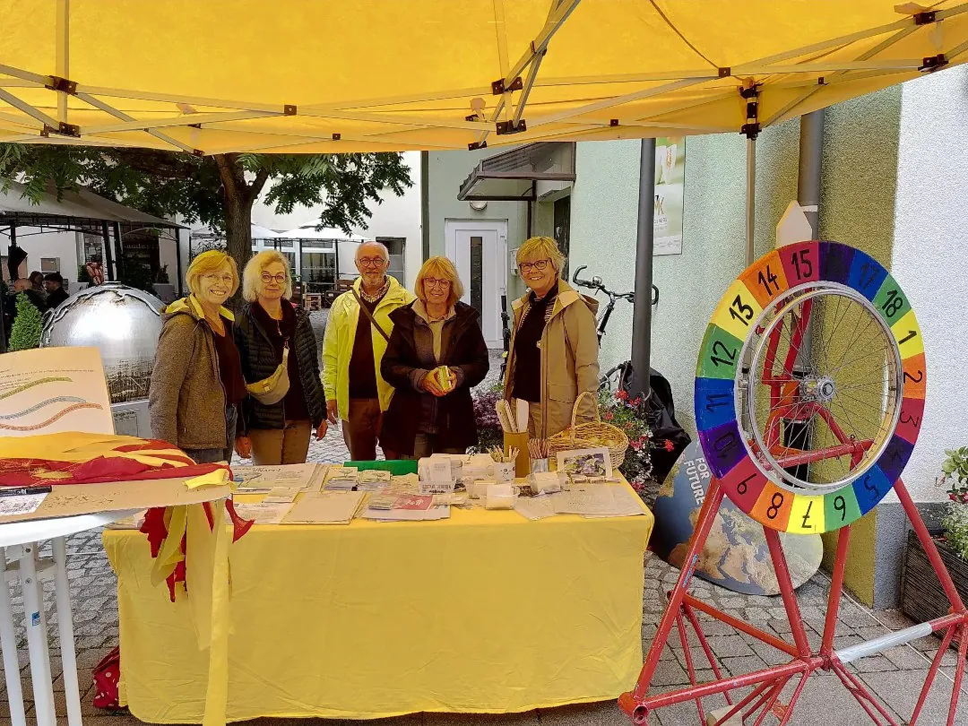 Fünf Personen stehen lächelnd hinter einem mit Informationsmaterial bedeckten Tisch unter einem gelben Zelt. In der Nähe steht ein buntes Glücksrad, und im Hintergrund sind Bäume und Gebäude zu sehen.