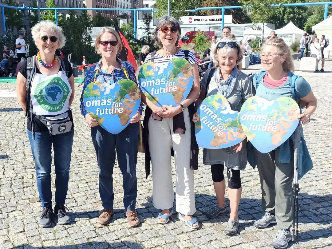 Fünf ältere Frauen stehen bei einer Kundgebung im Freien zusammen, lächeln und halten große Ausschnitte in Form der Erde mit dem Text "omas for future". Die sonnige Veranstaltung zeigt Stände und Menschen im Hintergrund.