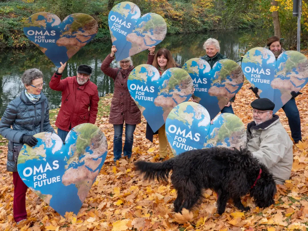Eine Gruppe von Senioren, die große herzförmige Schilder mit einer Weltkarte und dem Text OMAS FÜR DIE ZUKUNFT halten, steht im Freien auf herabgefallenen Herbstblättern und lächelt gemeinsam in der Nähe eines Teiches. Ein schwarzer Hund ist ebenfalls im Vordergrund zu sehen.