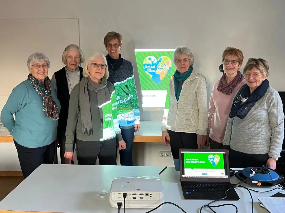 Sieben ältere Frauen stehen gemeinsam in einem hellen Raum vor einem projizierten Bild mit einem Herz und der Weltkugel. Ein Laptop und ein Projektor stehen auf einem weißen Tisch im Vordergrund. Die Frauen lächeln in die Kamera.