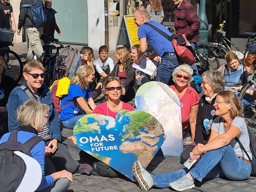 Eine Gruppe älterer Erwachsener sitzt im Kreis auf einer Kopfsteinpflasterstraße, lächelt und hält große herzförmige Schilder mit Erdbildern und dem Text OMAS FÜR DIE ZUKUNFT. Im Hintergrund sind Fahrräder und weitere Menschen zu sehen.